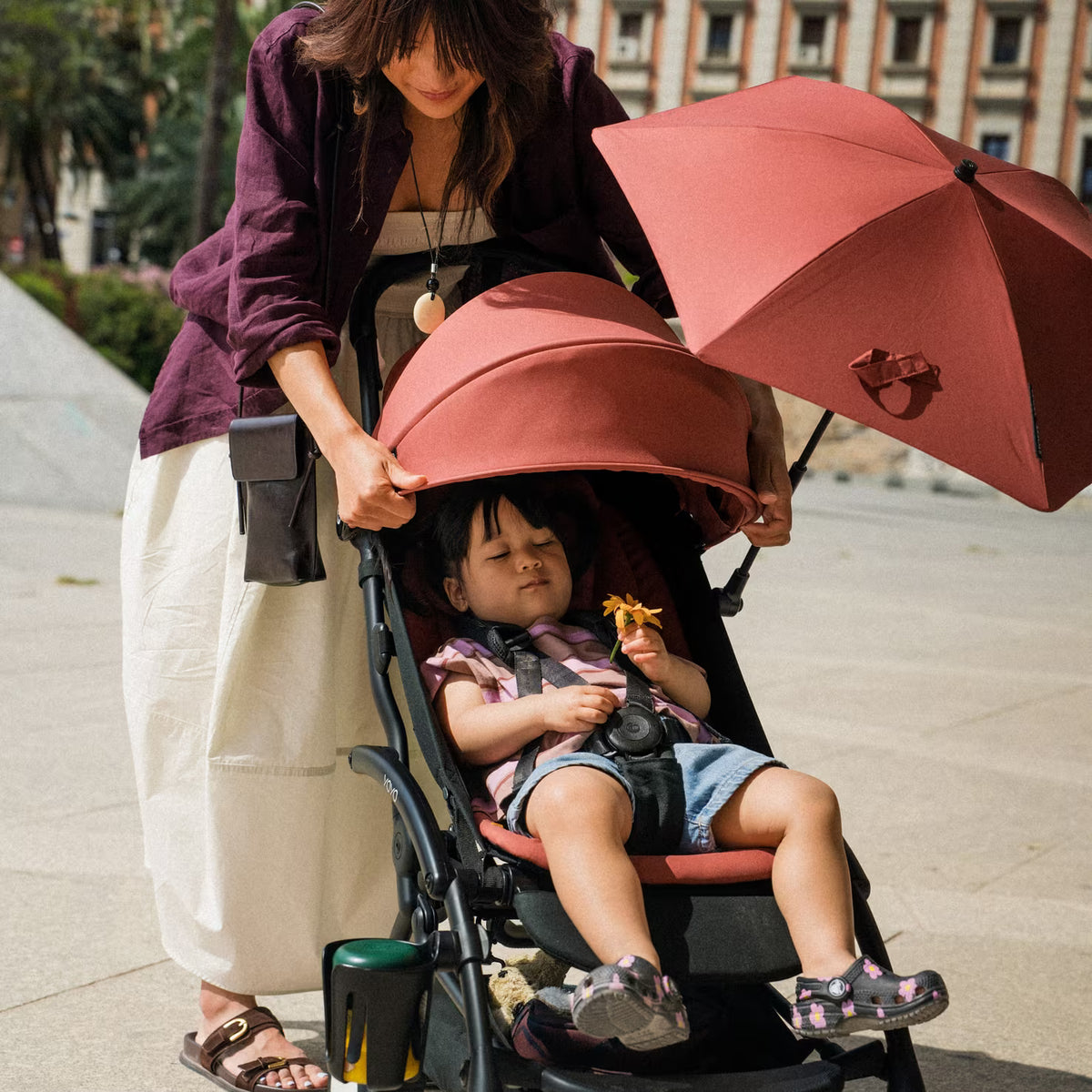 A woman pushes a stroller with a child inside on a sunny day. The stroller has a large dusty rose pink sun canopy and matching pink inner shade, with a black frame.