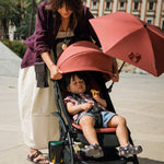 A woman pushes a stroller with a child inside on a sunny day. The stroller has a large dusty rose pink sun canopy and matching pink inner shade, with a black frame.