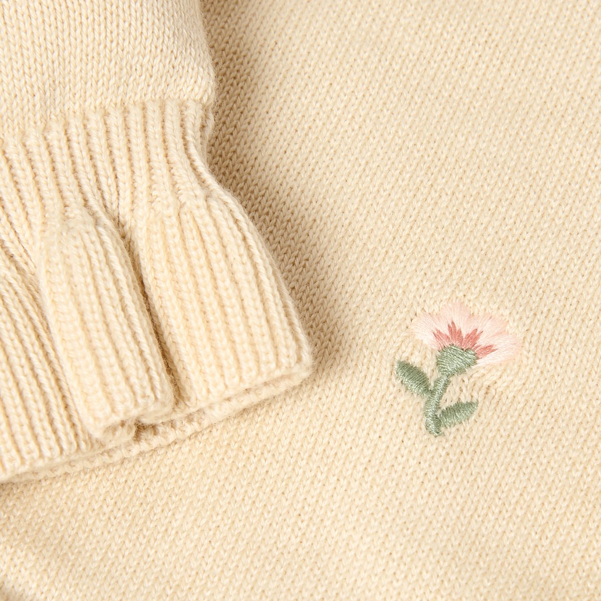 Close-up of a cream-colored knit sweater showing a ribbed cuff and a small embroidered pink flower with green leaves on the chest.