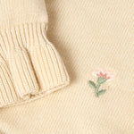 Close-up of a cream-colored knit sweater showing a ribbed cuff and a small embroidered pink flower with green leaves on the chest.