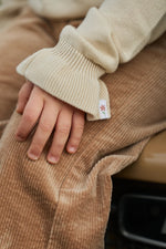 Close-up of a child's hand resting on brown corduroy pants, wearing a cream ribbed knit sweater with a small embroidered flower logo on the cuff.