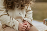 A close-up lifestyle shot of a child wearing a cream ribbed knit sweater with a small embroidered flower on the chest, paired with brown corduroy trousers, sitting outdoors.